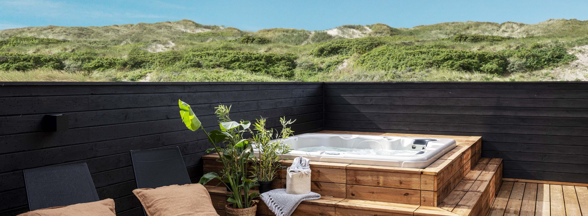 Sommerhus-terrasse i træ med udendørs spa og strandstole i hjørnet samt sandklitter og blå himmel i baggrunden.